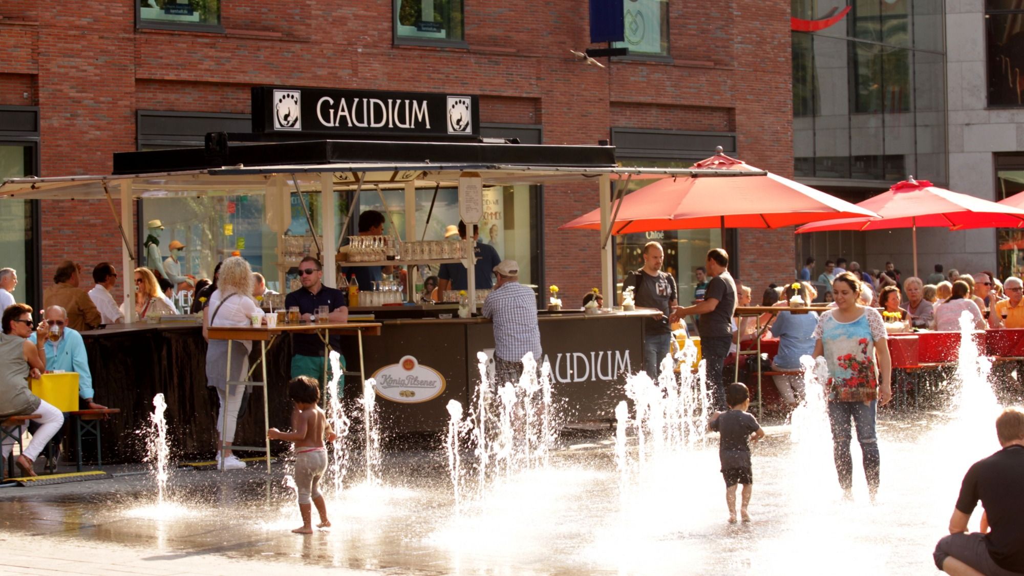 GAUDIUM-Getränkestand mit Logo auf einem sommerlichen Stadtfest, Gäste an Stehtischen und Kinder am Wasserspiel
